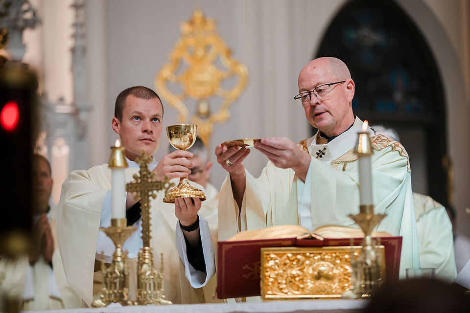 A bishop and a deacon in white vestments perform Catholic Mass at the altar, holding a chalice and host amid candles and gold decor.