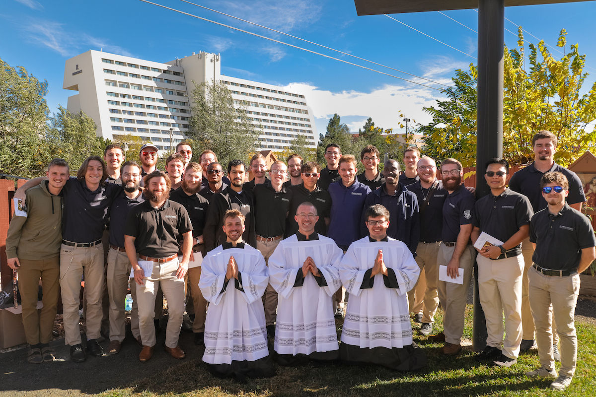 Large group of men posing outdoors in front of a hotel, with three in white robes kneeling in front under a bright blue sky.