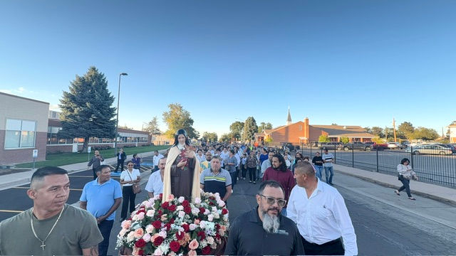 Crowd carries a flower-covered Virgin Mary statue down a street at dusk, with church and school buildings in the background.