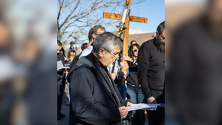 Man in black robe reads from a paper at an outdoor religious gathering, with a large wooden cross and crowd behind him.