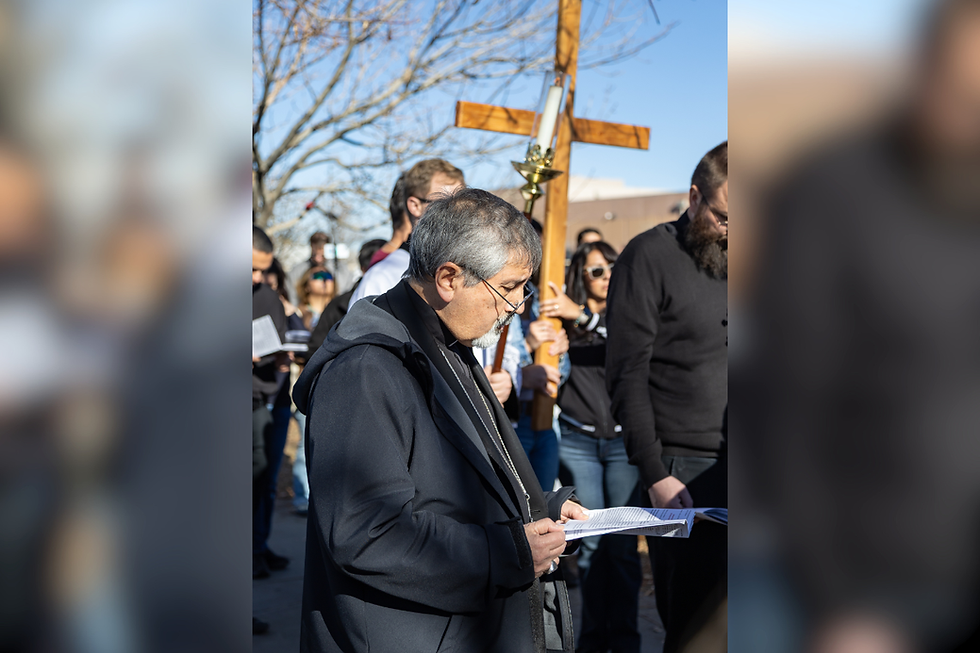 Man in black robe reads from a paper at an outdoor religious gathering, with a large wooden cross and crowd behind him.