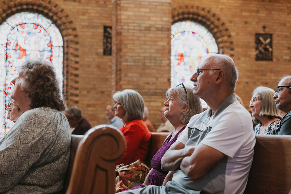 Older adults sit in a church pew listening quietly, with stained-glass windows and warm brick walls in the background.