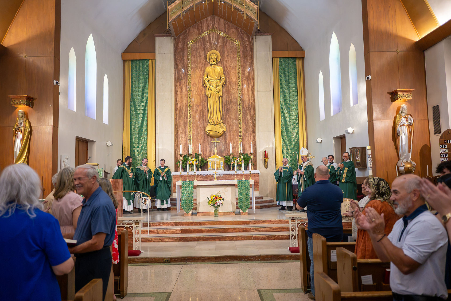Congregation applauds priests in green vestments inside a church sanctuary with a large golden statue above the altar.