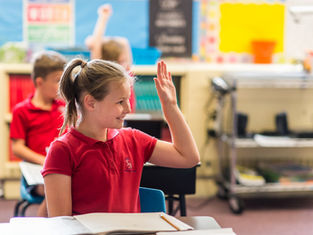 Girl in a red polo raises her hand in a bright classroom, smiling at her desk while other students sit blurred behind her.