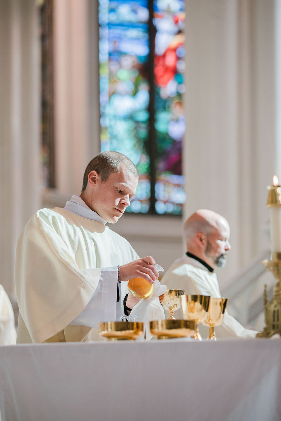 Deacon in white vestments pours wine into a chalice at a church altar, with stained glass glowing in the background.
