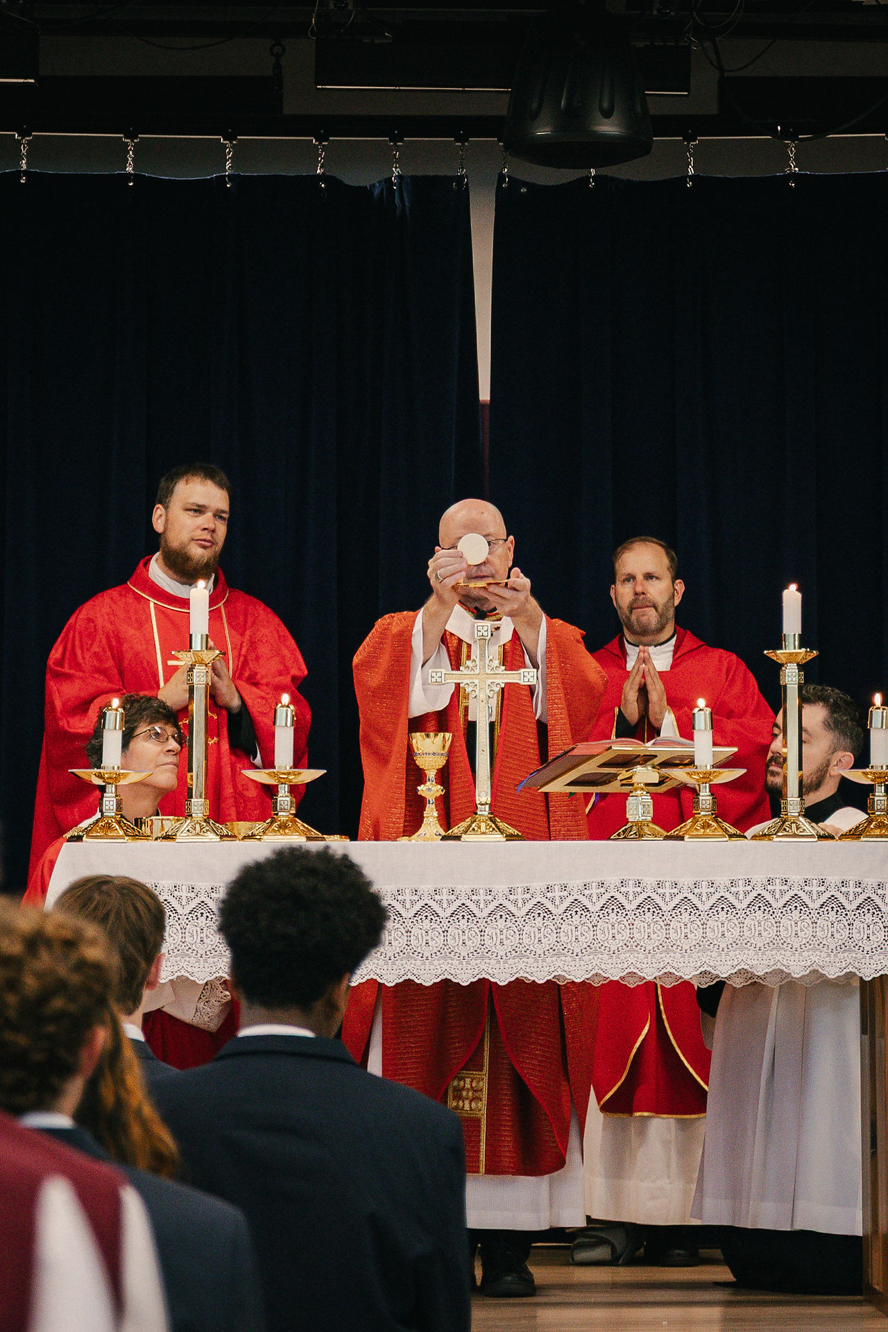 Priests in red vestments celebrate communion at a candlelit altar before dark curtains, with worshippers watching quietly.