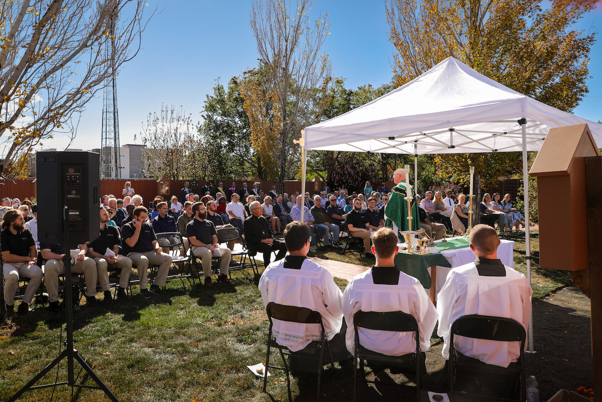 Outdoor Catholic service under a white tent, with a priest at the altar, altar servers seated, and a large congregation in autumn sun.