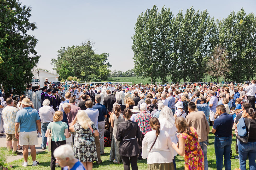 Large crowd gathered outdoors for a ceremony in a grassy field, with clergy and attendees facing a central speaker under tall trees