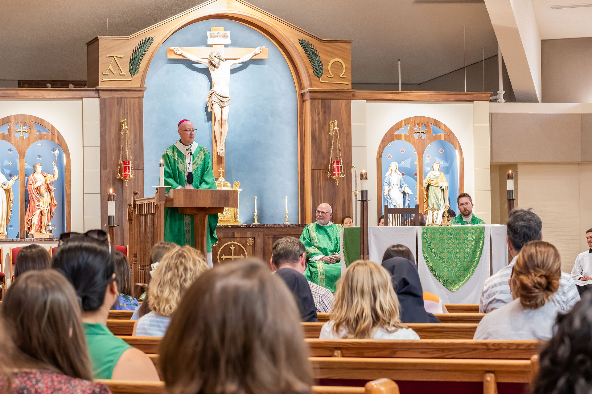 Priest in green vestments speaks at lectern before crucifix in a church, with seated congregation and altar candles behind him.