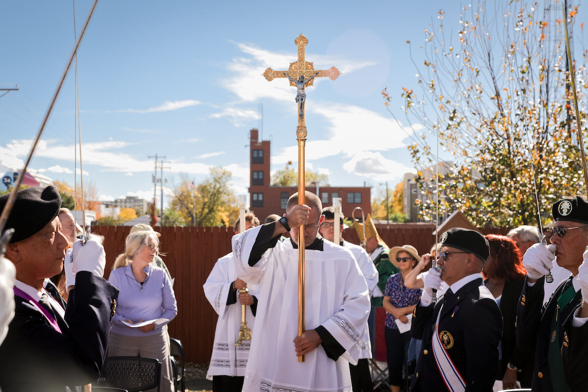 Priest in white robe holds a gold crucifix during an outdoor ceremony, flanked by uniformed veterans saluting under a blue sky.