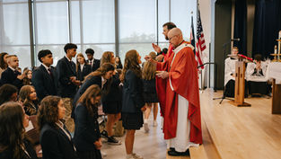 Priest in red vestments gives communion to kneeling students in a bright chapel with flags and altar servers in the background