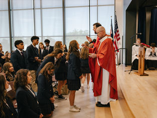 Priest in red vestments gives communion to kneeling students in a bright chapel with flags and altar servers in the background