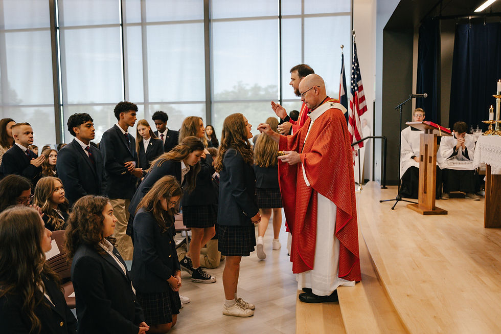 Priest in red vestments gives communion to kneeling students in a bright chapel with flags and altar servers in the background