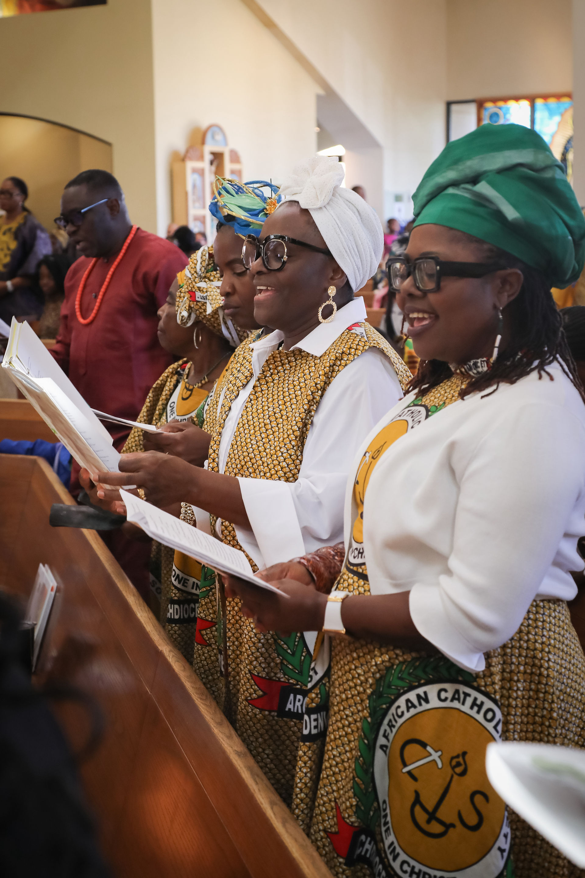 Women in colorful headwraps and patterned church robes sing from hymn sheets in a bright church, smiling in worship.