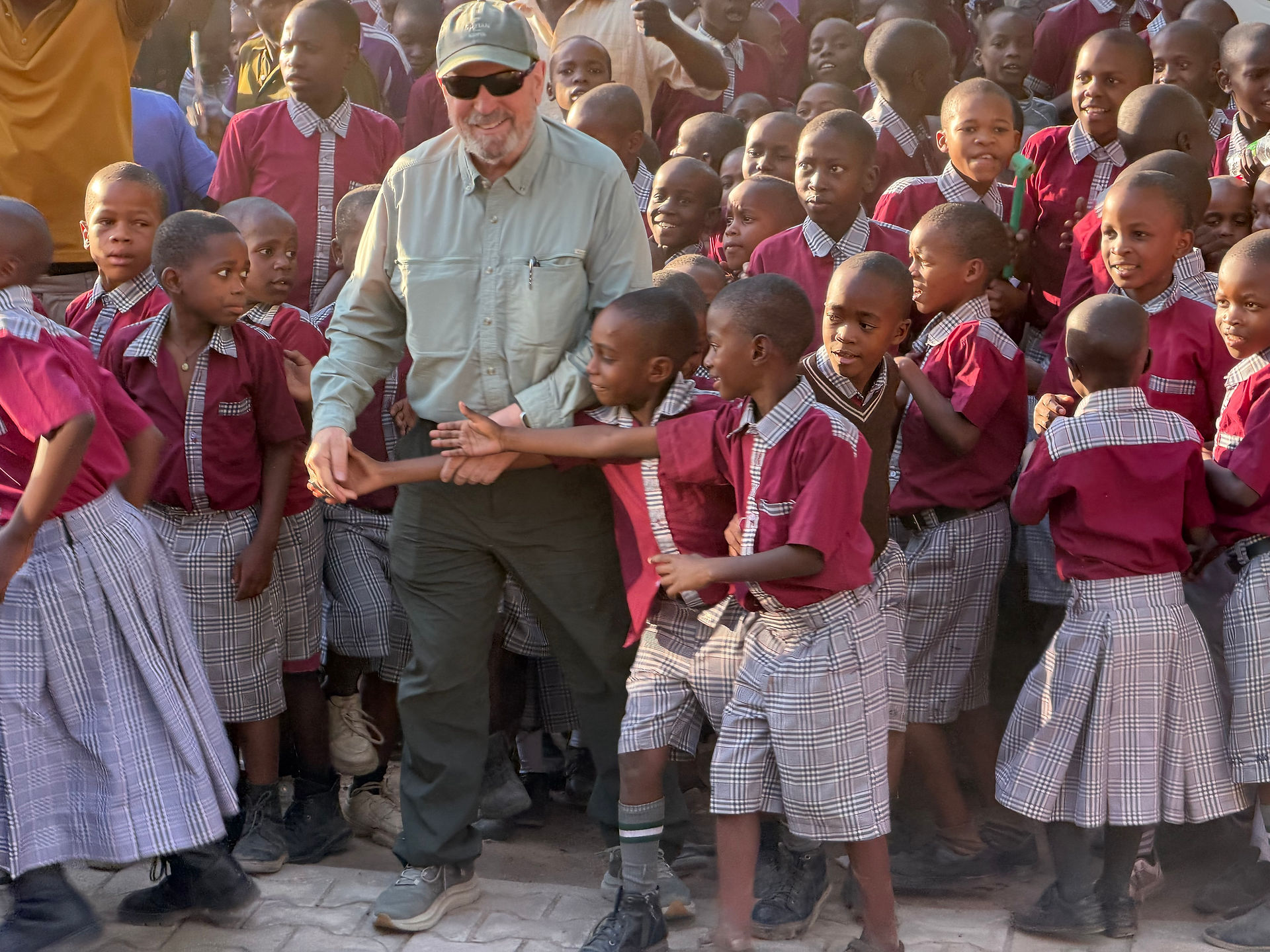 Smiling man in cap and sunglasses stands amid cheering schoolchildren in maroon uniforms, outdoors in warm sunlight.