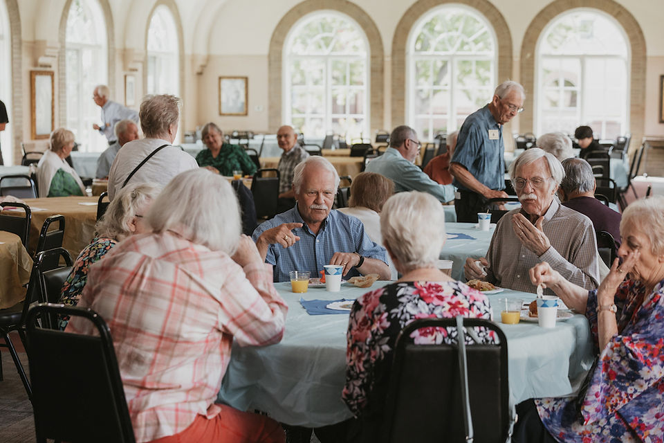 Older adults chat and eat at round tables in a bright banquet hall with tall arched windows, a relaxed social gathering