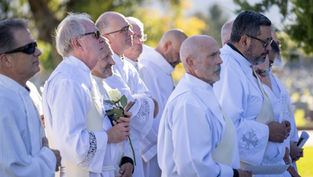 Men in white robes stand outdoors in sunlight at a solemn ceremony, one holding a white rose.