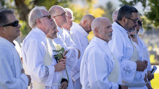 Men in white robes stand outdoors in sunlight at a solemn ceremony, one holding a white rose.