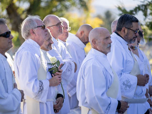 Men in white robes stand outdoors in sunlight at a solemn ceremony, one holding a white rose.