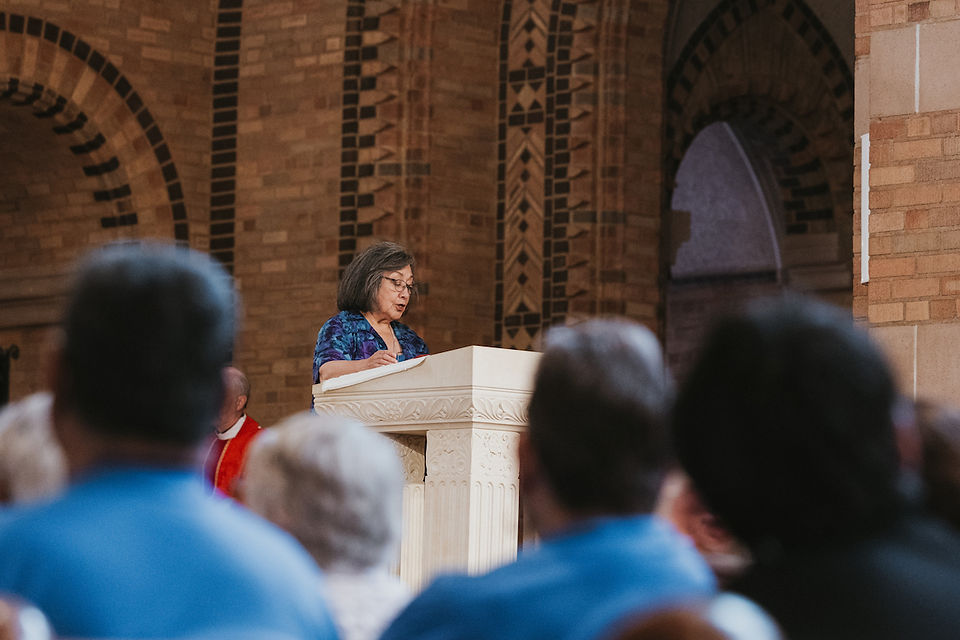 Woman speaks at a carved podium in a brick church, seen from a blurred audience under arched windows.