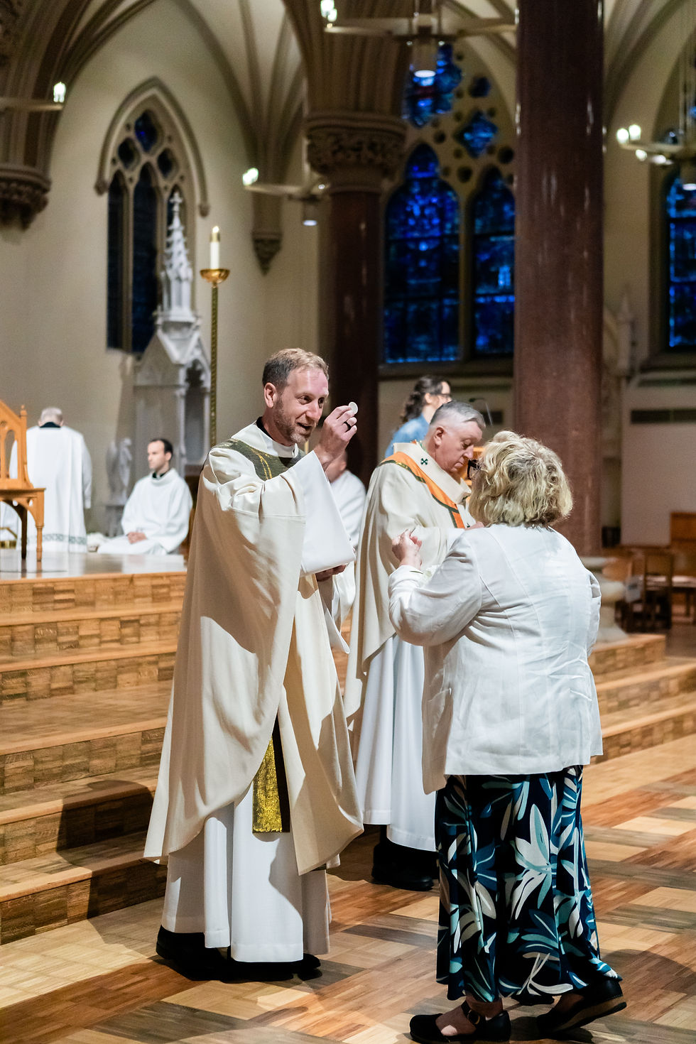 Priests in white vestments give communion to a woman inside a cathedral with blue stained glass and warm wooden floors.