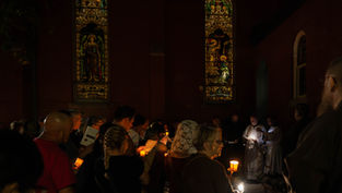 Candlelit congregation sings outside a dark church beneath stained-glass windows, with clergy reading from sheets.