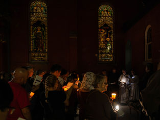 Candlelit congregation sings outside a dark church beneath stained-glass windows, with clergy reading from sheets.