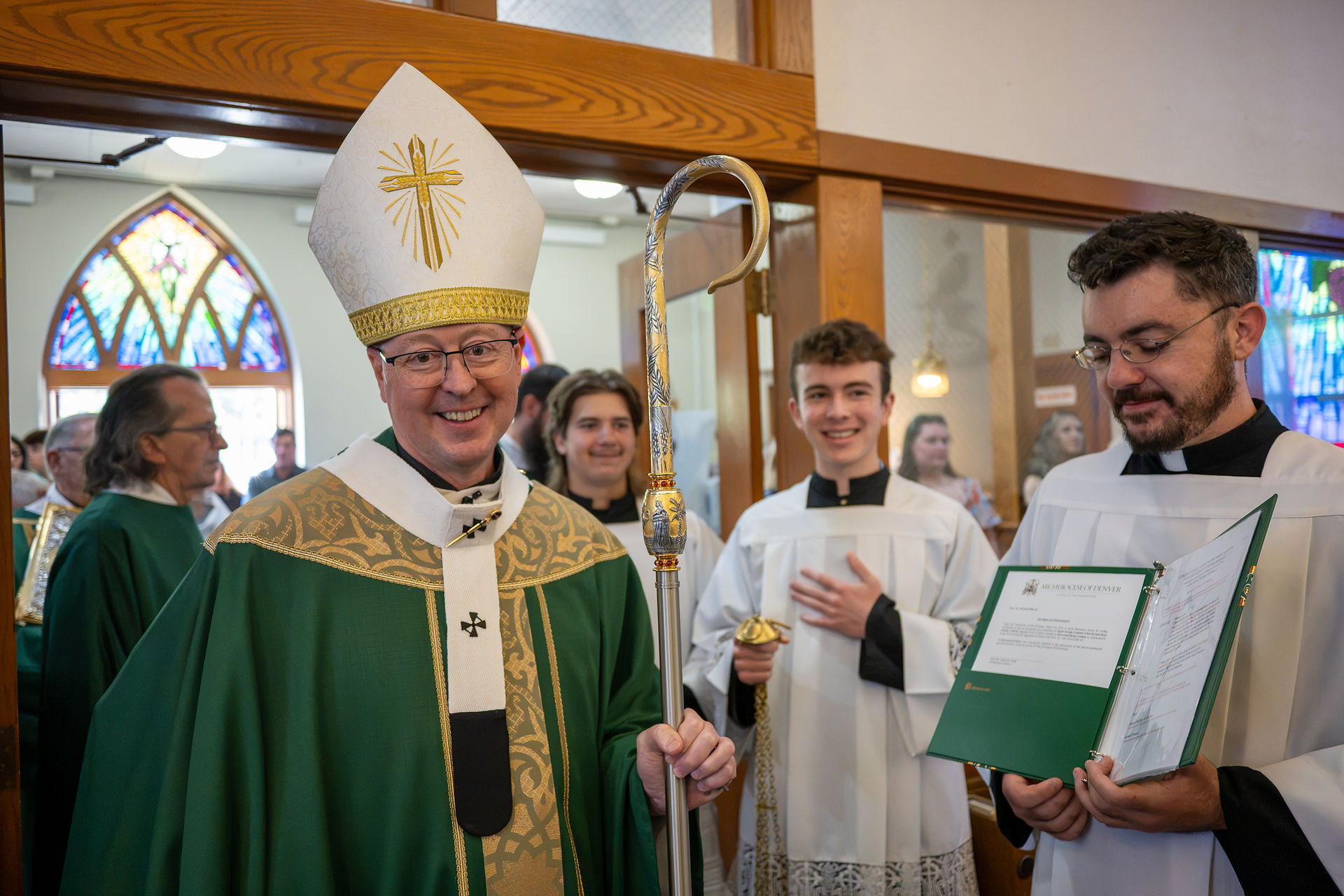 Smiling bishop in green vestments and mitre holds a crozier with altar servers in white robes inside a church with stained glass.