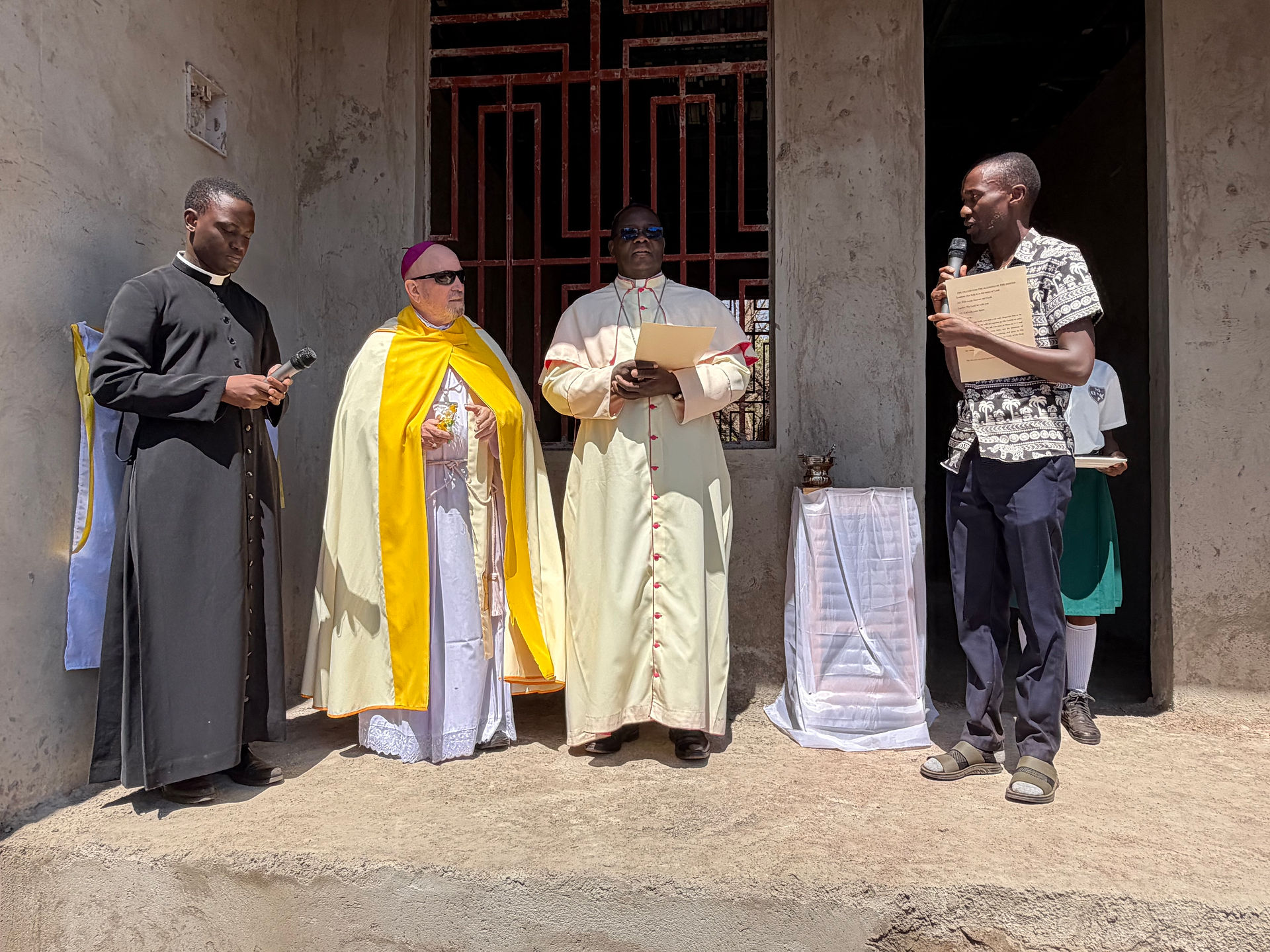 Clergy and laymen stand outside a church wall, one speaking into a microphone and others holding papers in bright sun.