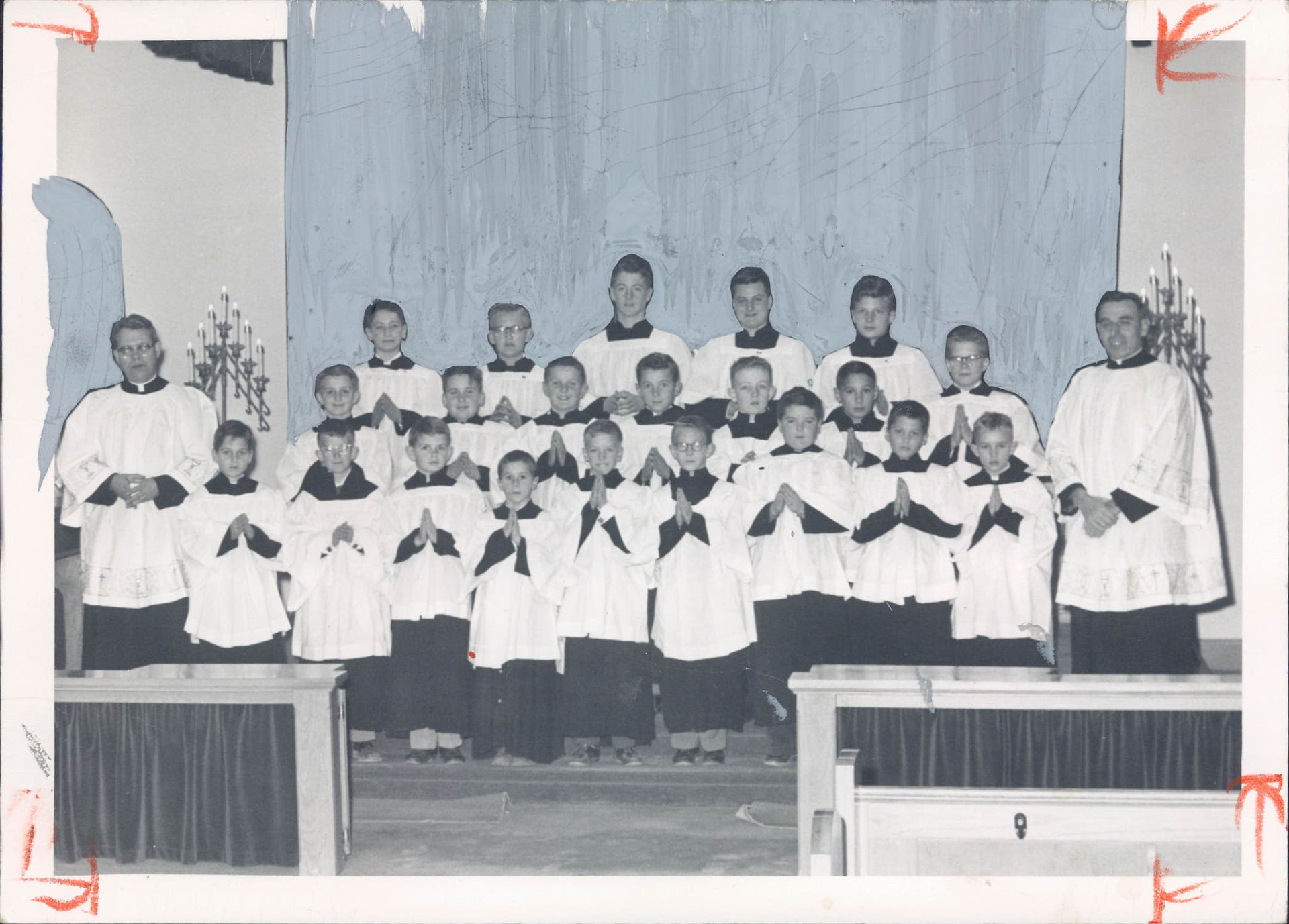 Boys’ church choir in white robes poses with two priests before a blue backdrop, hands folded in prayer.