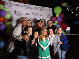 Smiling group poses at a Catholic Charities event backdrop, framed by colorful balloons under an EXIT sign.