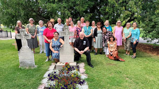 Smiling group poses at a cemetery by stone crosses and flowered grave, under a large green tree on a sunny day