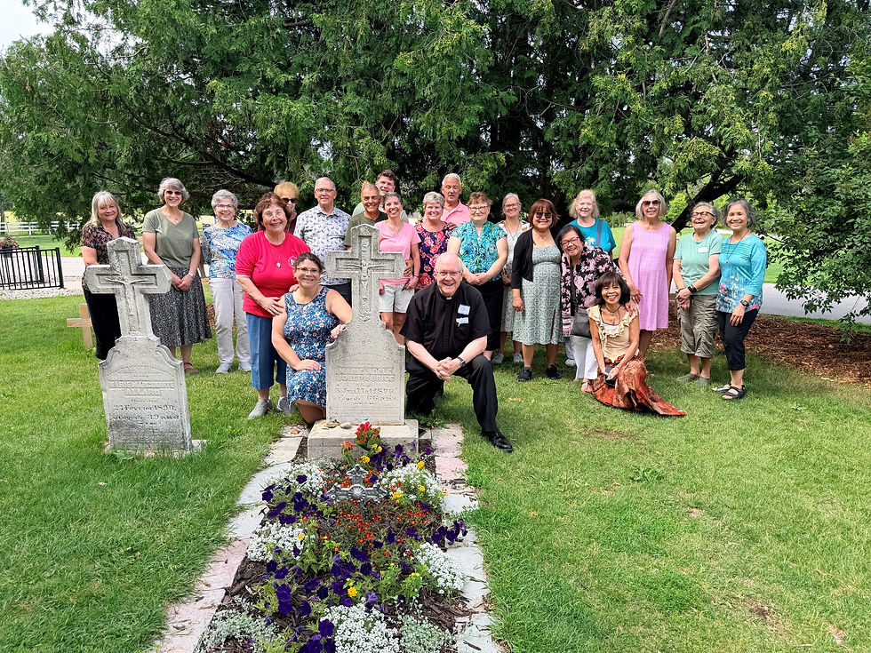 Smiling group poses at a cemetery by stone crosses and flowered grave, under a large green tree on a sunny day