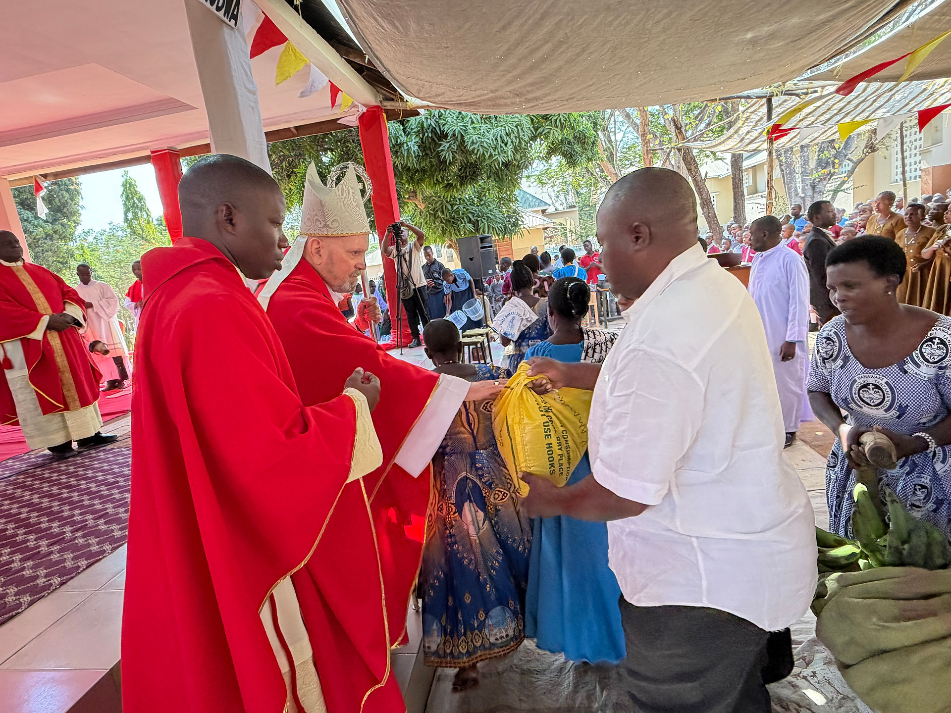 Priests in red robes hand a yellow sack to a man in white at an outdoor church ceremony under bunting, with crowd watching