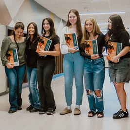 Six smiling students in a school hallway hold Quincy yearbooks, standing by classroom doors.