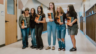Six smiling students in a school hallway hold Quincy yearbooks, standing by classroom doors.