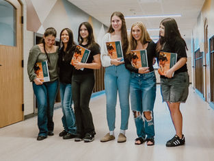 Six smiling students in a school hallway hold Quincy yearbooks, standing by classroom doors.