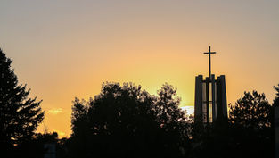 Silhouetted church bell tower with cross above trees at sunset, glowing orange sky and calm evening mood.