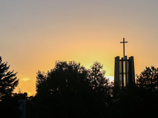 Silhouetted church bell tower with cross above trees at sunset, glowing orange sky and calm evening mood.