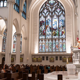 Bishop in ornate robes reads at a pulpit in a bright Gothic church, with seated congregation and stained-glass windows behind him.