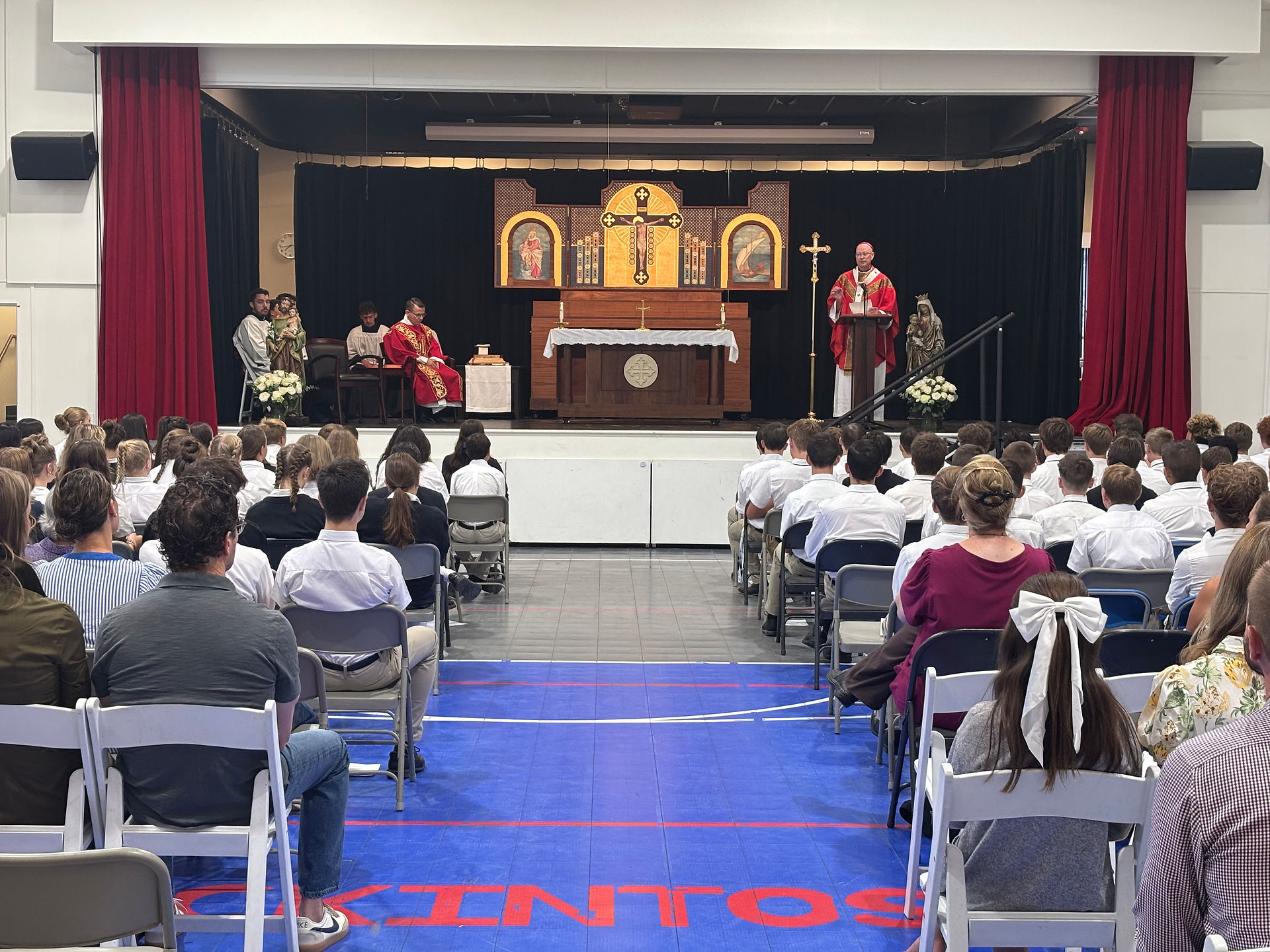 Priest in red vestments leads a church service on a school stage, with students and staff seated facing the altar and cross.