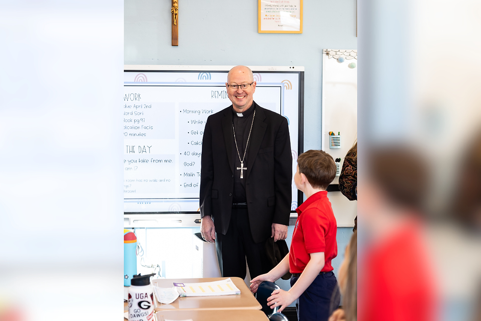 Smiling priest in black suit and cross stands in a classroom beside a boy in a red shirt, with lesson notes on a whiteboard.