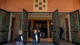 Children in school uniforms walk into a brick church through open decorated doors, with adults ahead in a calm, reverent scene.