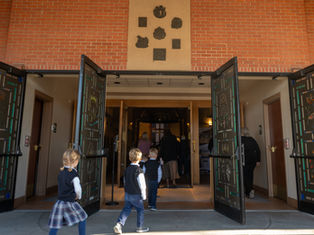 Children in school uniforms walk into a brick church through open decorated doors, with adults ahead in a calm, reverent scene.