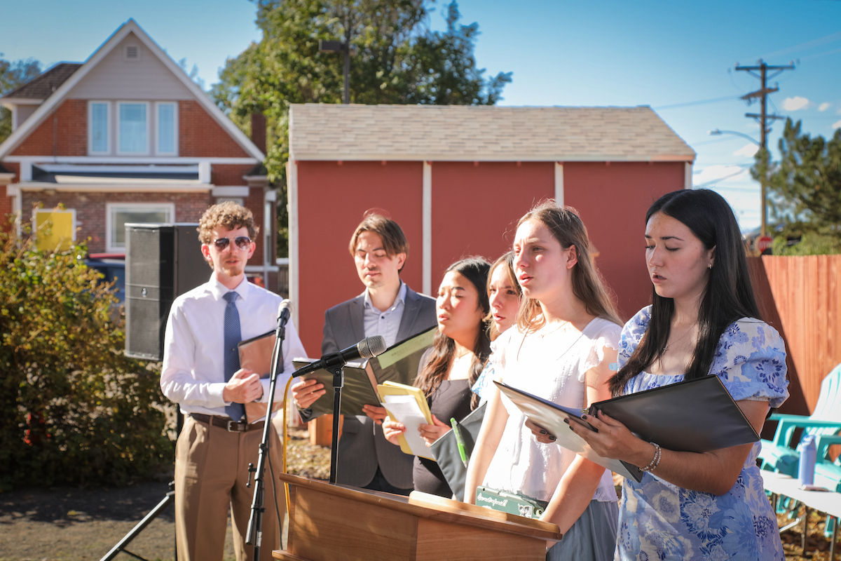 Six young adults sing from folders at microphones in a sunny backyard, with houses and a red shed behind them.