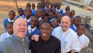 Smiling schoolboys in blue uniforms pose with three clergy outdoors in a sunny courtyard.