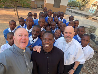 Smiling schoolboys in blue uniforms pose with three clergy outdoors in a sunny courtyard.