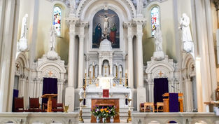 Ornate church altar with crucifix painting, statues, stained glass, purple drapes, candles, and flower arrangement in a quiet sanctuary