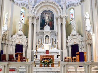 Ornate church altar with crucifix painting, statues, stained glass, purple drapes, candles, and flower arrangement in a quiet sanctuary