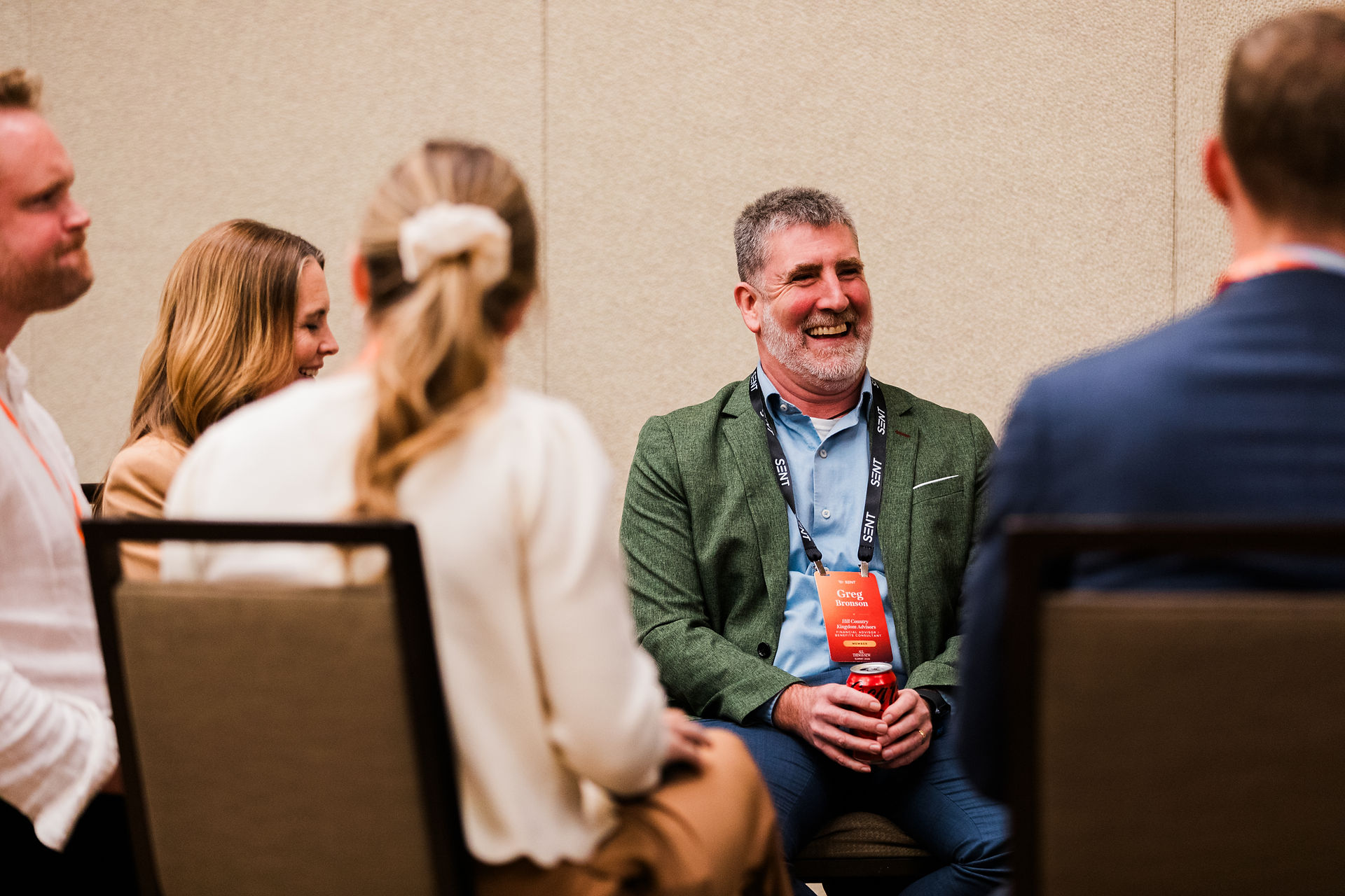 Smiling man in green blazer with Greg Bronson badge chats with attendees at a conference.